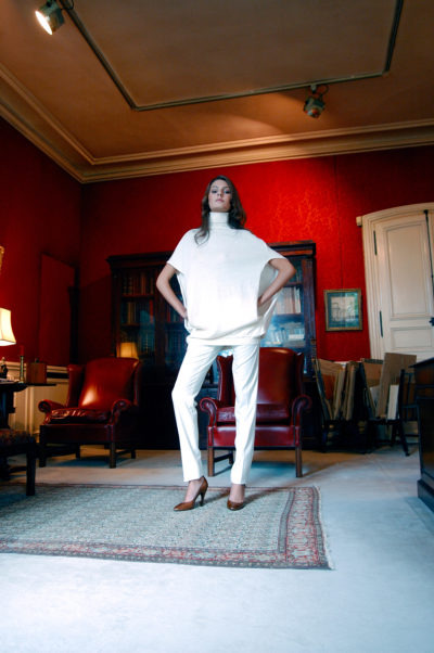 Une mannequin vêtue d'une tenue entièrement blanche, composée d'un haut ample et d'un pantalon, se tient debout, les mains sur les hanches, dans une pièce élégante aux murs d'un rouge intense, aux meubles classiques et à un tapis clair. Le photographe de mode Thomas Duclert capture la scène avec des fauteuils en cuir, des étagères et des œuvres d'art encadrées.