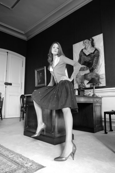 Photographie en noir et blanc d'une mannequin assise au bord d'un bureau dans une élégante pièce parisienne. Elle porte un cardigan, un chemisier et une jupe, une jambe croisée sur l'autre. La pièce, est ornée d'œuvres d'art encadrées sur les murs, d'un bureau en bois, de portes blanches fermées et d'un tapis.