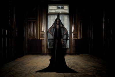 Une personne se tient dans une pièce sombre et opulente, aux boiseries et aux grandes fenêtres. Elle est vêtue d'une longue robe noire fluide avec un voile transparent couvrant son visage, projetant une ombre étrange sur le sol. Les détails palatiaux de la pièce créent une atmosphère envoûtante et dramatique qui rappelle la mode parisienne de Thomas Duclert.