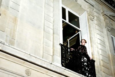 Une mannequin en robe noire se tient sur un balcon en fer forgé, regardant par une grande fenêtre d'un élégant immeuble en pierre à Paris. Le bâtiment est doté de hautes fenêtres, de volets blancs et de détails architecturaux ornementaux. La scène est illuminée par la lumière, soulignant sa présence.