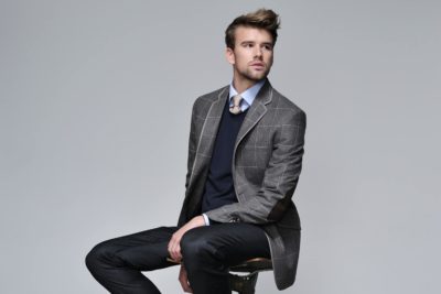 Un mannequin aux cheveux bruns courts et à la barbe est assis sur un tabouret sur un fond uni, rappelant le studio d'un photographe parisien. Il porte un blazer à carreaux gris sur un pull foncé et une chemise à col bleu clair, associés à un pantalon foncé. Il regarde pensivement au loin.