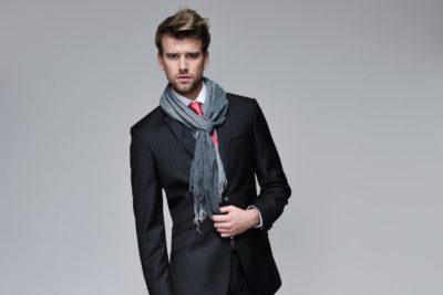 Un mannequin vêtu d'un costume sombre à rayures et d'une chemise blanche avec une cravate rouge, accessoirisée d'un foulard gris, incarne la mode. Debout sur un fond gris uni, il regarde l'objectif avec assurance tout en ajustant le foulard d'une main. Le photographe capture son style soigné sans effort.