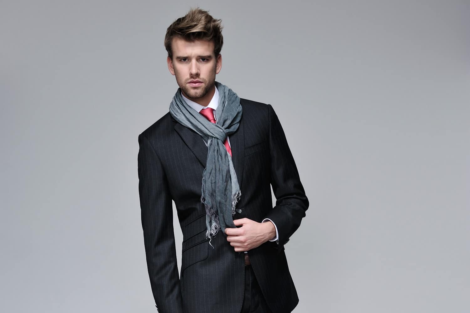 Un mannequin vêtu d'un costume sombre à rayures et d'une chemise blanche avec une cravate rouge, accessoirisée d'un foulard gris, incarne la mode. Debout sur un fond gris uni, il regarde l'objectif avec assurance tout en ajustant le foulard d'une main. Le photographe capture son style soigné sans effort.