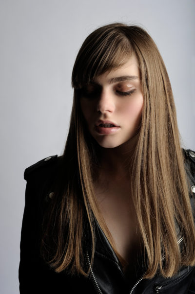 Une mannequin aux longs cheveux bruns raides et à la frange se tient debout sur un fond gris. Elle porte une veste en cuir noir et regarde vers le bas, les yeux fermés, dégageant un air de calme et de sérénité. Le photographe capture cet instant serein, faisant écho à l'essence chic de la mode parisienne.