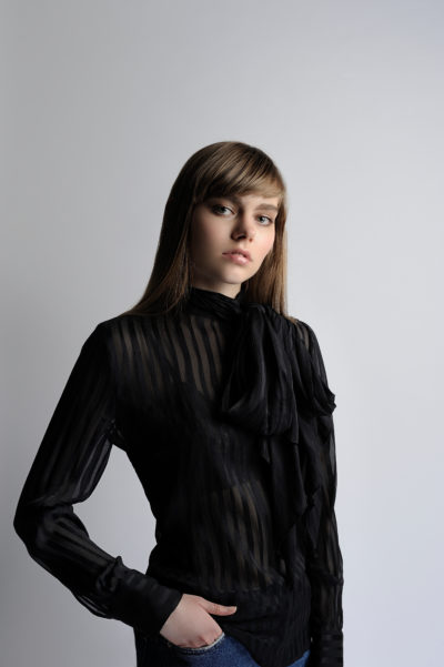 Une personne aux cheveux longs et raides, coiffée d'une frange, se tient debout sur un fond gris uni. Elle porte une blouse noire transparente à rayures verticales, une cravate oversize au cou et a une main dans la poche de son jean. Évoquant la mode parisienne à la Thomas Duclert, elle regarde directement la caméra.