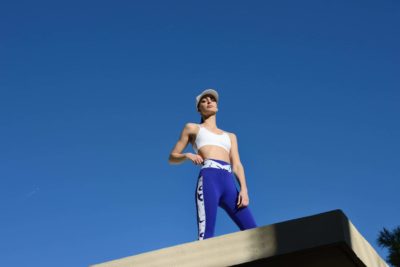 Une mannequin en tenue de sport se tient debout, confiante, sur une structure en béton devant un ciel bleu clair. Elle porte un soutien-gorge de sport blanc, des leggings bleus avec des accents blancs et une casquette blanche, le regard levé et détourné de l'objectif. Photographiée par Thomas Duclert, l'image respire la force et la détermination.