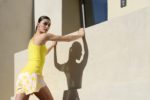 Une mannequin en débardeur jaune et short à motifs s'appuie contre un mur beige, peut-être en train de s'étirer ou de faire de l'exercice. Son ombre est projetée sur le mur alors qu'elle se tient à l'extérieur près d'un bâtiment moderne avec une fenêtre, le tout magnifiquement capturé par le photographe Thomas Duclert à Paris.