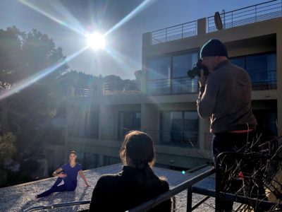 Un photographe, coiffé d'un chapeau, prend en photo une personne posant sur un toit de Paris, illuminé par le soleil éclatant. Une autre personne, dos à l'objectif, observe la scène. En arrière-plan, on aperçoit un immeuble moderne aux larges fenêtres, immortalisant Thomas Duclert photographe au travail.