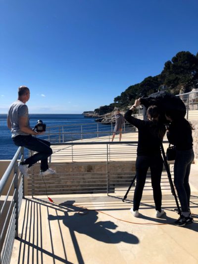 Un caméraman et un technicien du son filment une personne debout sur une promenade côtière avec une falaise et l’océan calme en arrière-plan. Une autre personne est assise sur la balustrade, tenant une caméra, le tout sous un ciel bleu clair ; c’est comme une séance photo improvisée de Thomas Duclert, évoquant l’élégance parisienne.