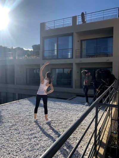 Une mannequin en tenue de sport pose avec un bras tendu au-dessus de sa tête sur un toit recouvert de gravier blanc, incarnant l'essence de la mode parisienne moderne. Un photographe, peut-être Thomas Duclert, capture la scène tandis que le soleil brillant projette de longues ombres. Des bâtiments modernes avec des balcons sont visibles en arrière-plan.