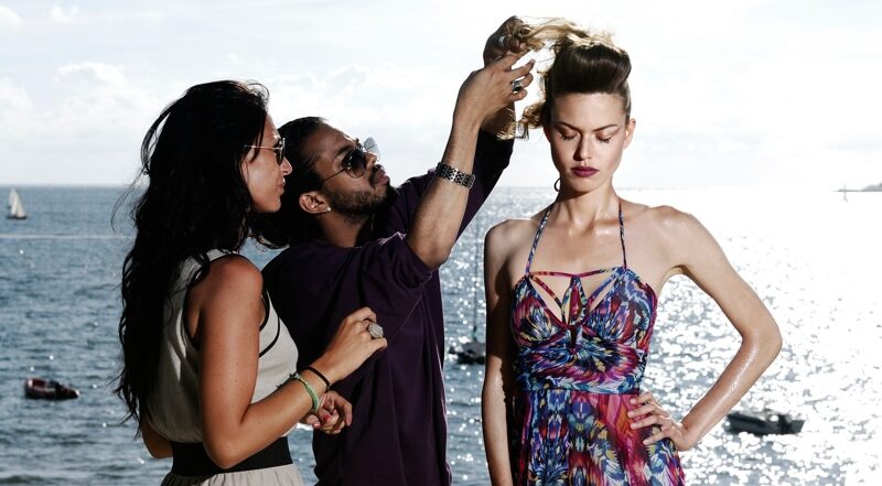 Une mannequin vêtue d'une robe colorée se tient au bord de l'océan tandis qu'on lui coiffe les cheveux et ajustent son look. Il utilise de la laque sur sa coiffure relevée, et l'autre femme lisse sa robe. L'arrière-plan ensoleillé présente un ciel clair et des voiliers sur l'eau.