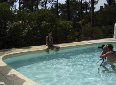 Une personne en maillot de bain noir est assise au bord d'une piscine, les pieds dans l'eau, tandis qu'une autre personne, peut-être Thomas Duclert, se tient debout dans la piscine et capture l'instant. La scène se déroule dans un décor de verdure luxuriante et d'arbres.