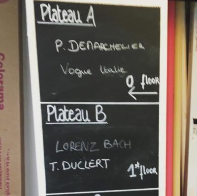 Un panneau en ardoise répertoriant les séries et les noms. Sous « Plateau A », on peut lire « P. Denarcheler, Vogue Italia, rez-de-chaussée ». Sous « Plateau B », on peut lire « Lorenz Bach, Thomas Duclert, premier étage ». L'arrière-plan présente des boîtes de dossiers sur des étagères, ce qui est parfait pour un décor de photographe de mode.