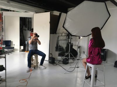 Le photographe Thomas Duclert capture des images d'une mannequin aux cheveux longs portant une robe rose brillante. La personne est assise élégamment sur un tabouret devant une grande lampe de studio avec un réflecteur parapluie blanc. Ce studio de photographie professionnel et lumineux, rappelant la mode parisienne, met en valeur divers équipements soigneusement disposés.