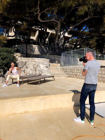 Un homme en t-shirt gris et en jean prend une photo avec un appareil photo. Il photographie un autre homme en débardeur blanc et en short qui est assis sur un banc et pose. Ils sont dehors par une journée ensoleillée à Paris, entourés de murs en pierre et d'arbres.