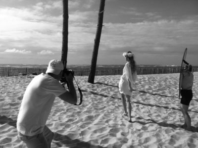 Photographie en noir et blanc du photographe Thomas Duclert et d'une mannequin debout sur une plage de sable. La mannequin porte une robe légère et un surfeur tenant une planche est visible à l'arrière-plan. Deux grands arbres sont également visibles sur la plage, évoquant le charme intemporel de l'art parisien au bord de la côte.