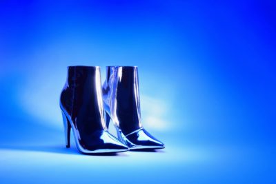 Une paire de bottines à talons hauts en métal argenté brillant, illuminées par un fond bleu dégradé vibrant. Habilement capturées par le photographe parisien Thomas Duclert, les bottes sont positionnées côte à côte, avec leurs bouts pointus et leur finition élégante reflétant la lumière.