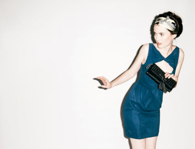 Une mannequin à la peau claire et aux cheveux foncés, enveloppée dans un foulard, porte une robe bleue sans manches. Elle tient une pochette noire et regarde sur le côté avec une expression surprise. Prise par le photographe Thomas Duclert, l'arrière-plan est blanc uni.