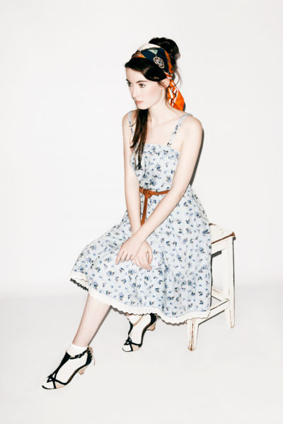 Une mannequin est assise sur un tabouret blanc sur un fond uni. Elle porte une robe bleu clair à motifs floraux ceinturée par une ceinture marron, des talons noirs et blancs et un foulard coloré noué autour de la tête. Elle regarde sur le côté avec une expression neutre, capturant la quintessence de la mode parisienne évoquant le style photographique de Thomas Duclert.