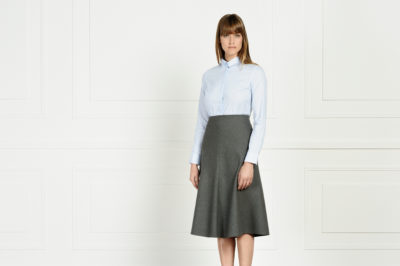Une mannequin aux cheveux bruns raides se tient debout devant un mur lambrissé blanc. Elle porte une chemise blanche boutonnée rentrée dans une jupe trapèze gris anthracite à taille haute qui lui arrive aux genoux. Imitant la mode parisienne, elle regarde directement la caméra avec une expression neutre, rappelant l'élégance discrète de Thomas Duclert.