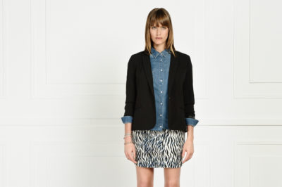Une mannequin aux cheveux longs se tient debout sur un fond neutre, à l'image de la mode parisienne. Elle porte une chemise en jean sous un blazer noir et une jupe à motifs noirs et blancs. Ses bras sont détendus le long de son corps et son expression neutre rappelle le style emblématique de Thomas Duclert.