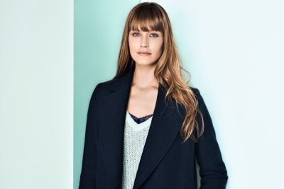 Une mannequin aux cheveux longs et raides et à la frange se tient debout sur un fond clair. Elle porte un manteau sombre sur un haut clair. Son expression est neutre alors qu'elle regarde directement l'objectif, dégageant une mode parisienne parfaitement capturée par le photographe.