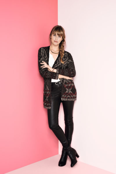 Une mannequin aux cheveux longs tressés sur une épaule est appuyée contre un mur divisé en deux moitiés roses et blanches. Elle est en mode mode, portant un cardigan à motifs noirs, un haut blanc, un pantalon en cuir noir et des bottines noires, le tout capturé avec brio par le photographe Thomas Duclert.
