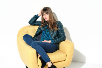 Une mannequin aux cheveux longs est assise confortablement sur une chaise jaune vif sur un fond blanc uni, ressemblant à une scène tout droit sortie de Paris. Elle porte une chemise à motifs bleu canard, une veste matelassée verte, un jean bleu et des bottines noires. Sa pose est détendue, avec un bras posé sur le dossier de la chaise et l'autre main touchant ses cheveux.