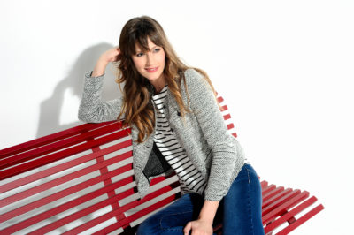 Une mannequin aux cheveux longs et bruns est assise sur un banc rouge sur fond blanc. Photographiée par Thomas Duclert, elle porte un blazer gris sur une chemise rayée noire et blanche et un jean bleu. Elle sourit et appuie sa tête sur sa main droite, qui repose sur le dossier du banc.