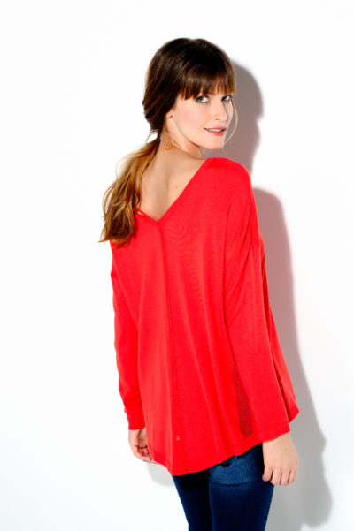 Une mannequin aux longs cheveux bruns se tient debout et regarde par-dessus son épaule, dans un style parisien. Elle porte un haut ample rouge avec un décolleté en V dans le dos et un jean bleu, rappelant le style intemporel de Thomas Duclert. Le fond est blanc uni.