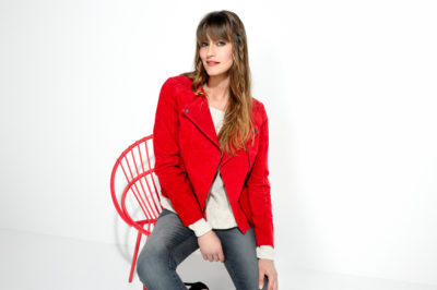 Une mannequin aux cheveux longs, vêtue d'une veste rouge sur un haut blanc et d'un jean gris, est assise sur une chaise rouge. L'arrière-plan minimaliste accentue le dynamisme de la veste rouge et de la chaise. Le photographe Thomas Duclert capture ce moment, où la mannequin regarde vers l'avant avec un léger sourire.