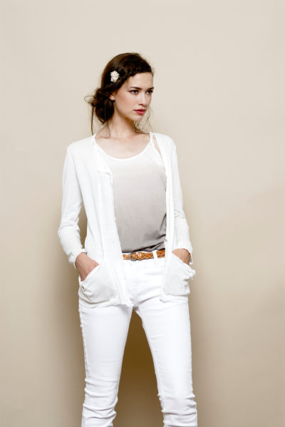 Une mannequin se tient debout sur un fond neutre, à l'image de la mode parisienne, vêtue d'un cardigan blanc sur un haut gris clair et d'un pantalon blanc. Les mains dans les poches de son cardigan et une ceinture marron serrée à la taille, elle incarne l'élégance associée à Thomas Duclert. Ses cheveux sont coiffés avec un petit accessoire sur le côté.