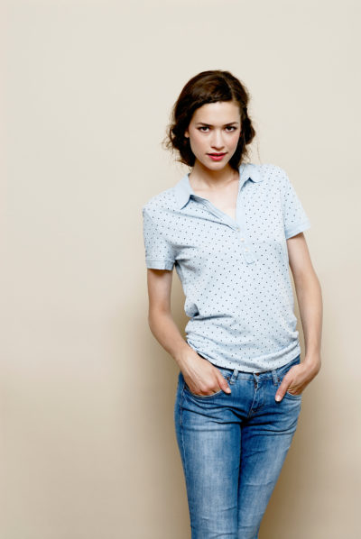Une mannequin aux cheveux bruns ondulés jusqu'aux épaules, portant une chemise à manches courtes à pois bleu clair et un jean bleu, pose dans son meilleur style sur un fond beige avec une main dans la poche et un léger sourire.