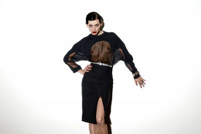 Une mannequin pose sur fond blanc dans un chemisier noir transparent à manches longues et une jupe noire taille haute fendue. Ses cheveux courts sont coiffés en arrière et elle accessoirise avec une ceinture, un bracelet et des bagues. Avec son rouge à lèvres rouge vif et son expression intense, elle respire la mode parisienne capturée par le photographe.