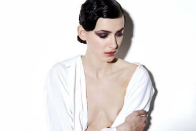 Une mannequin aux cheveux noirs lissés en arrière et au maquillage dramatique, notamment un fard à paupières violet et des lèvres rouges, regarde vers le bas, l'air contemplatif. Vêtue d'un vêtement blanc partiellement ouvert, exposant ses épaules et le haut de sa poitrine sur un fond blanc uni, elle évoque l'essence de la mode parisienne.