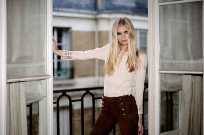 Une mannequin aux longs cheveux blonds se tient sur un balcon à Paris, appuyée contre les portes vitrées ouvertes à cadre blanc. Elle porte un chemisier beige avec des détails de boutons sur les poignets et un pantalon taille haute marron. L'arrière-plan, capturé par le photographe Thomas Duclert, montre un bâtiment avec une façade en briques et des fenêtres.