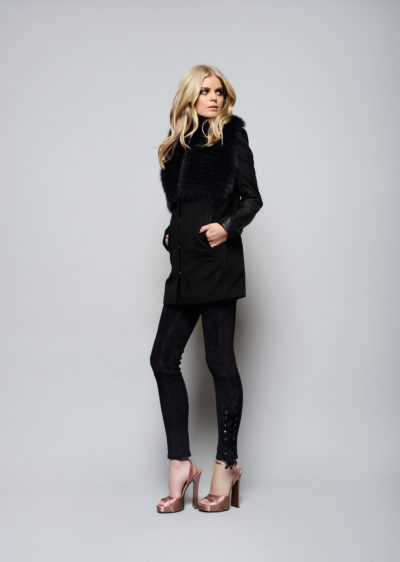 Une mannequin aux longs cheveux blonds se tient debout sur un fond gris uni, vêtue d'un manteau à col de fourrure noir, de leggings noirs et de chaussures à talons hauts métalliques. Avec une expression sérieuse et posée avec une main sur la hanche, elle incarne la haute couture parisienne.
