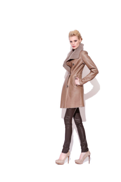Une personne aux cheveux blonds courts porte un élégant manteau croisé beige avec un large col bordé de fourrure. Elle porte un pantalon slim foncé et des chaussures beiges à talons hauts, posant parfaitement avec une main sur la hanche sur un fond blanc uni, capturé avec brio par le photographe Thomas Duclert.