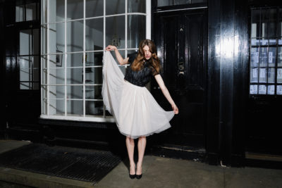 Une mannequin se tient sur un trottoir la nuit, soulevant d'une main l'ourlet de sa jupe en tulle blanc. Elle porte un haut noir et des talons hauts noirs, incarnant la dernière mode. Derrière elle se trouve un bâtiment avec de grandes fenêtres à cadre blanc et une porte noire, capturant l'élégance à travers l'objectif du photographe Thomas Duclert.