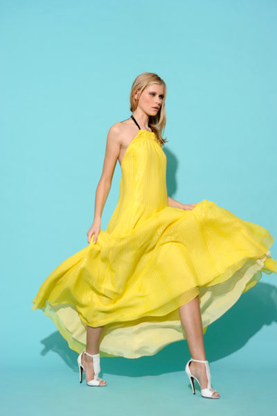 Une mannequin porte une robe fluide jaune vif à licou sur un fond bleu clair. Elle a les cheveux clairs et marche gracieusement, tenant les bords de sa robe qui s'enroule autour d'elle. Imitant la mode parisienne, elle porte également des sandales blanches à talons hauts, rappelant les créations de Thomas Duclert.