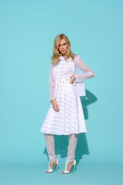 Une mannequin aux cheveux blonds se tient debout sur un fond bleu clair, vêtue d'une chemise blanche à manches longues à motifs, d'une jupe mi-longue, de leggings transparents et de talons hauts blancs. Elle tient un petit sac blanc et regarde directement l'objectif avec toute la prestance d'un photographe de mode parisien.