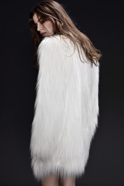 Une mannequin aux longs cheveux bruns, vêtue d'un manteau de fourrure blanc, se tient debout sur un fond sombre, légèrement détournée de l'objectif, la tête tournée sur le côté. La texture du manteau de fourrure est douce et moelleuse, contrastant avec le fond sombre, une scène qui rappelle la mode parisienne capturée par Thomas Duclert.