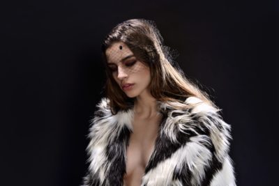 Une mannequin porte un manteau de fourrure noir et blanc et un voile noir sur le visage. Elle se tient debout sur un fond sombre, le regard baissé avec une expression sérieuse. Ses longs cheveux bruns tombent sur ses épaules, capturant l'essence de la mode parisienne à travers l'objectif du photographe.