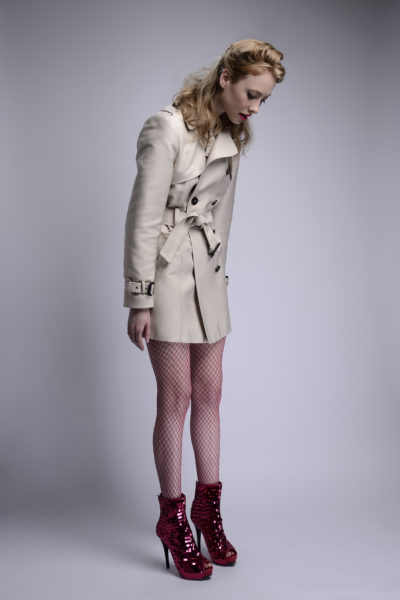 Une mannequin aux cheveux blonds se tient debout sur un fond uni, le regard baissé. Elle porte un trench-coat beige clair, des bas résille rouges et des bottines à talons hauts rouges brillants. Ses cheveux sont coiffés en vagues lâches, incarnant la mode capturée par le photographe Thomas Duclert.