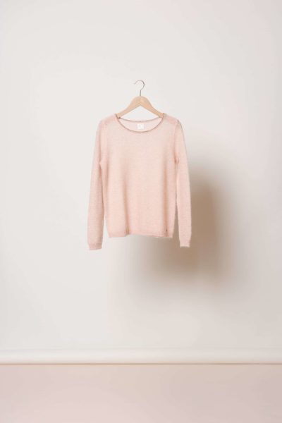 Un pull rose clair à manches longues de Thomas Duclert est suspendu à un cintre en bois devant un mur blanc uni. Ce pull, qui incarne la mode parisienne, présente une encolure ronde et un design simple avec une texture douce.