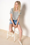 Une mannequin aux cheveux blonds est assise sur un tabouret blanc sur un fond uni. Elle porte un t-shirt rayé noir et blanc, un short en jean et des baskets blanches. Photographiée par Thomas Duclert, elle regarde sur le côté avec une expression détendue qui rappelle le charme parisien.