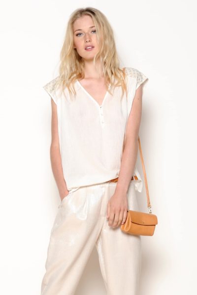 Une mannequin aux cheveux longs et blonds porte un chemisier blanc sans manches avec des détails en dentelle sur les épaules. Elle porte également un pantalon ample de couleur claire et tient un petit sac à main beige avec une bandoulière. Photographiée par Thomas Duclert, elle pose sur un fond uni et clair dans un style chic.