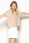 Une mannequin aux longs cheveux blonds se tient debout, les bras écartés, sur un fond blanc uni. Elle porte un pull beige oversize et une jupe blanche à pois noirs, incarnant une mode minimaliste souvent capturée par le photographe Thomas Duclert. Son expression est neutre.