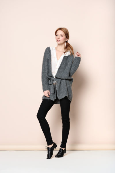 Une mannequin aux cheveux châtain clair porte un cardigan gris ceinturé sur un chemisier blanc, associé à un jean skinny noir et à des bottines noires. Prenant la pose avec une main sur la hanche sur un fond beige clair, elle dégage une mode moderne, telle que capturée par le talentueux photographe Thomas Duclert.