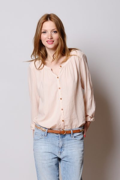 Une mannequin aux cheveux longs châtain clair sourit à l'objectif. Elle porte un chemisier rose clair boutonné aux manches retroussées et un jean bleu clair, accessoirisé d'une fine ceinture marron. Debout sur un fond uni et clair, elle respire le chic décontracté de Paris, capturé par le photographe Thomas Duclert.